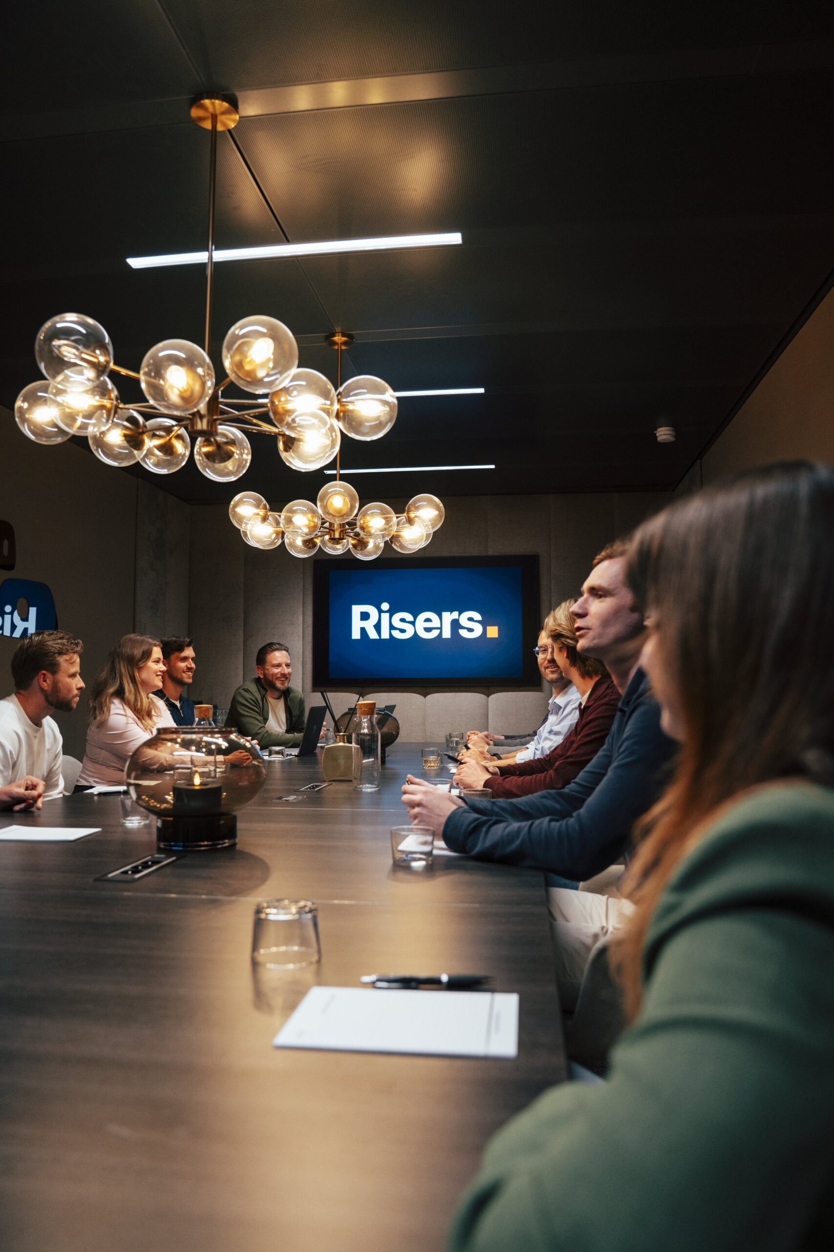 Risers-team in boardroom-gesprek met Risers-logo op scherm