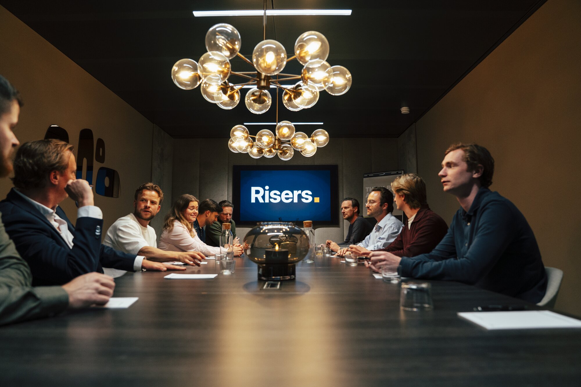 Volledig Risers-team rond boardroom-tafel, Risers-logo centraal op scherm